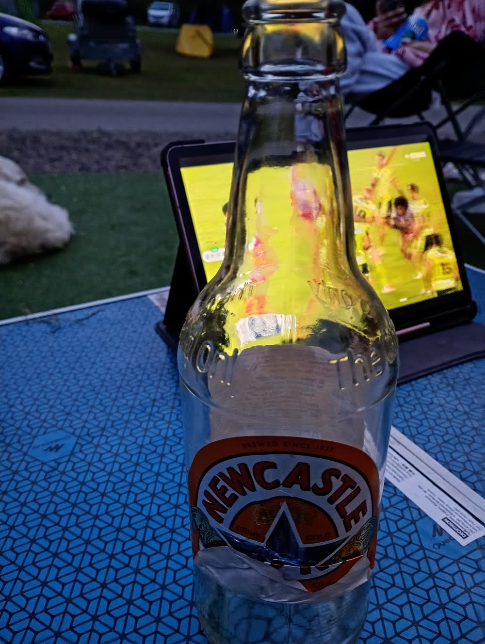 A Leigh rugby match streaming on a tablet beside a Newcastle Brown at the campsite